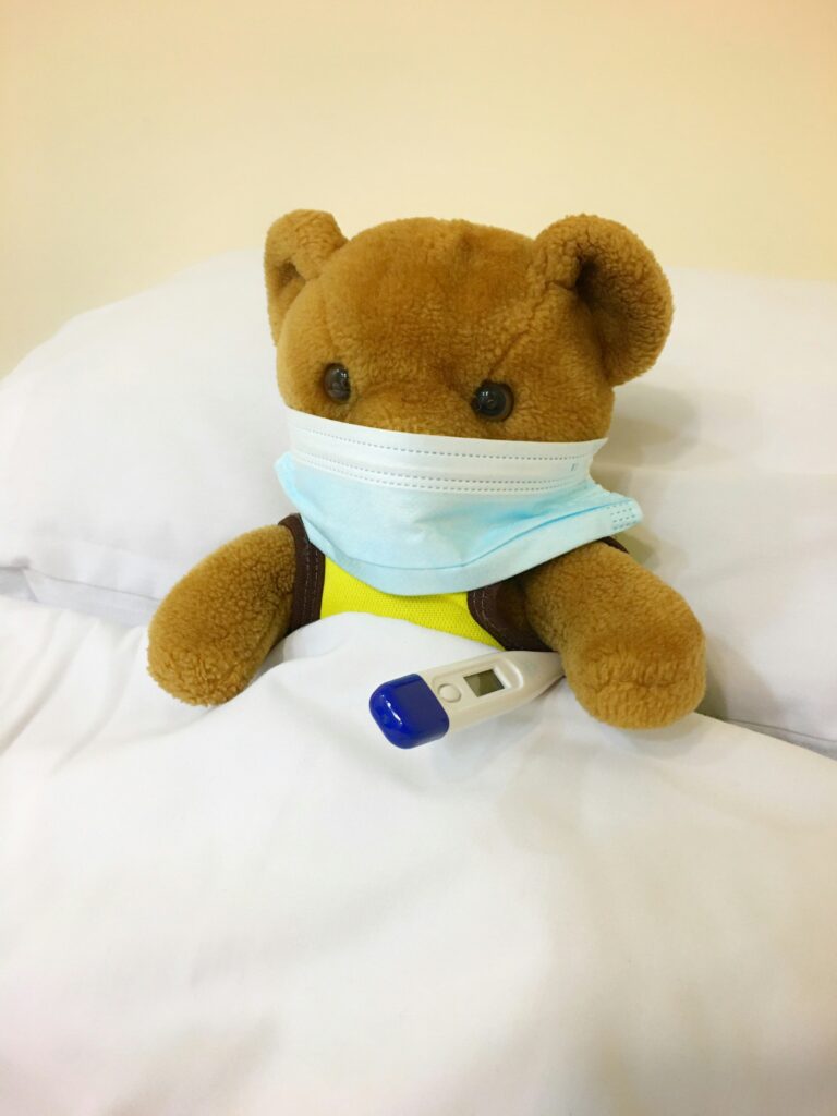 Teddibär mit Maske und Fieberthermometer zugedeckt sitzend im Bett